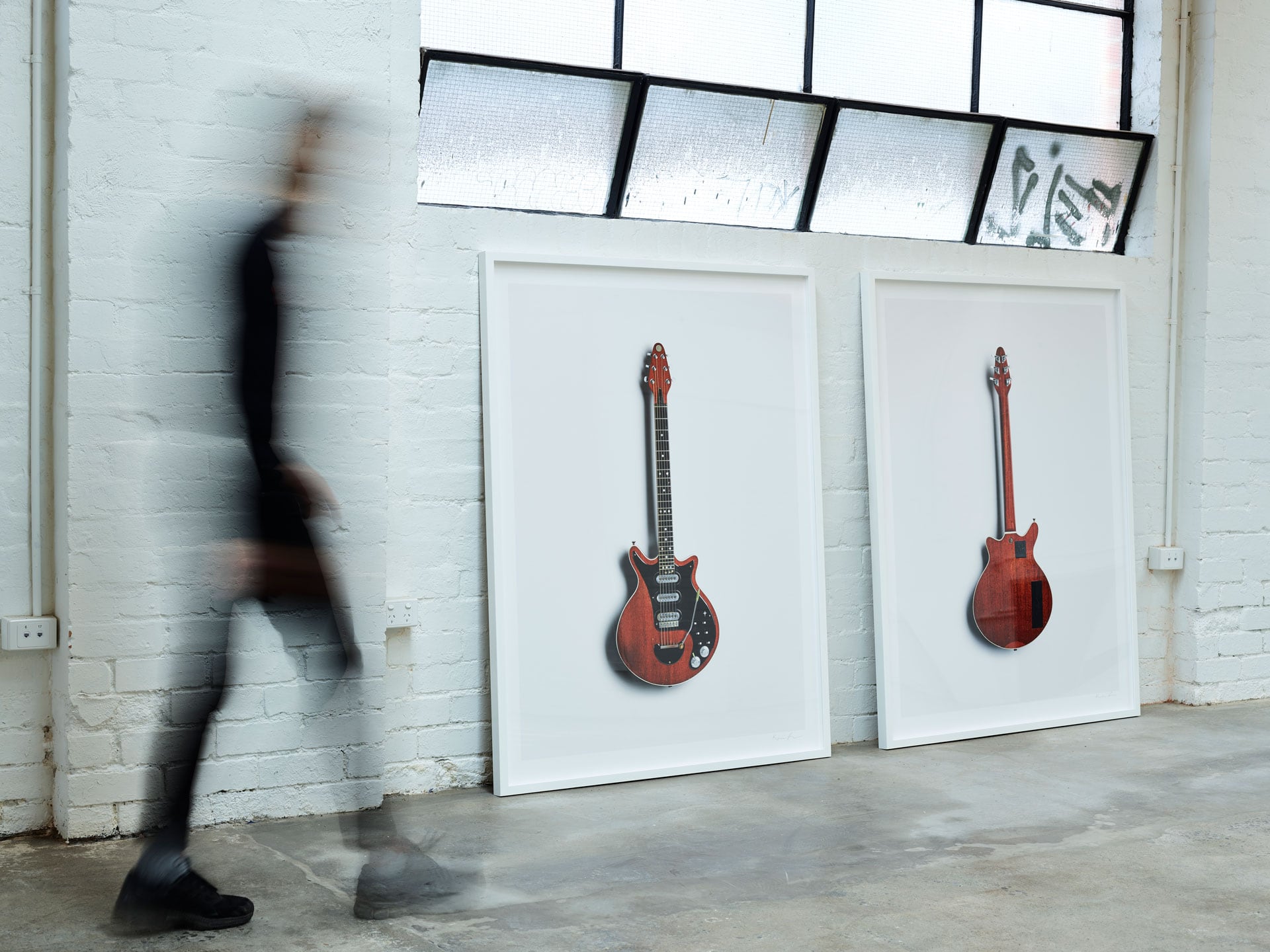 White framed images of guitar front and back are leant against a while brick wall. A person moves on the left, blurred by their motion.