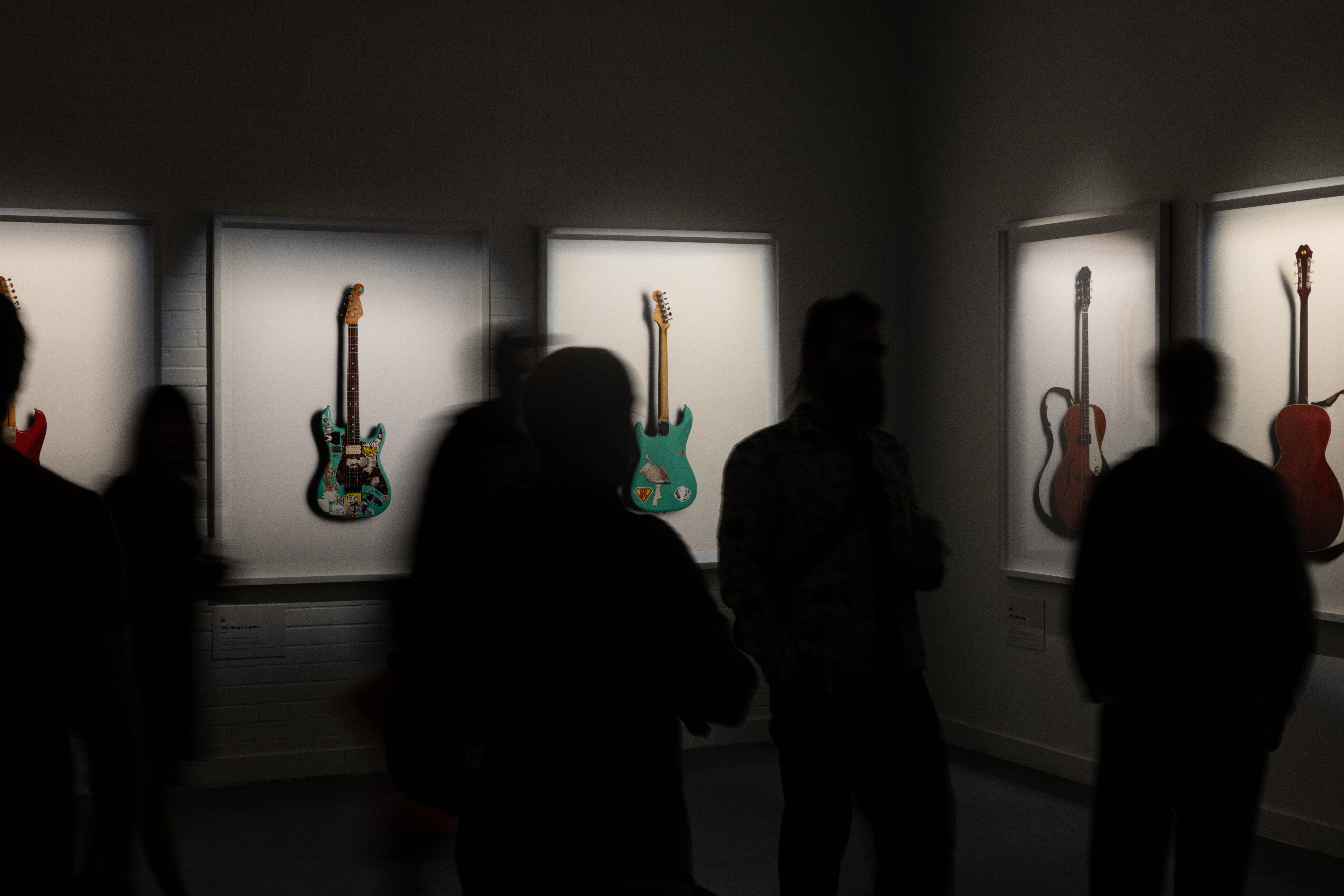 Guitars images in white frames are displayed on a white brick wall and lit from above. The back of people viewing them can be seen in the foreground.