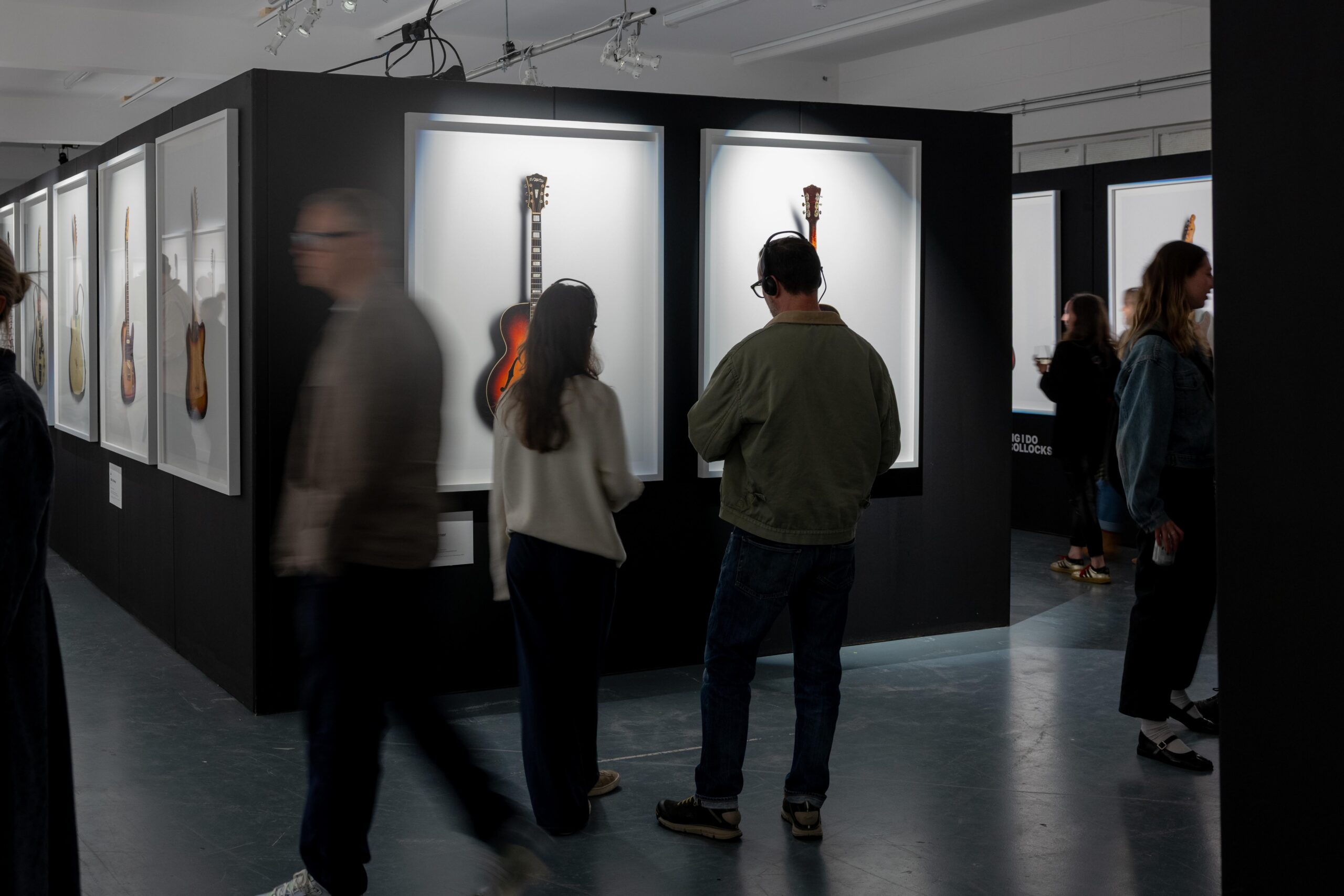 Two people stand in front of an image of a guitar in a white frame, obscuring the guitar from view. A person walking past appears blurred by their. movement.