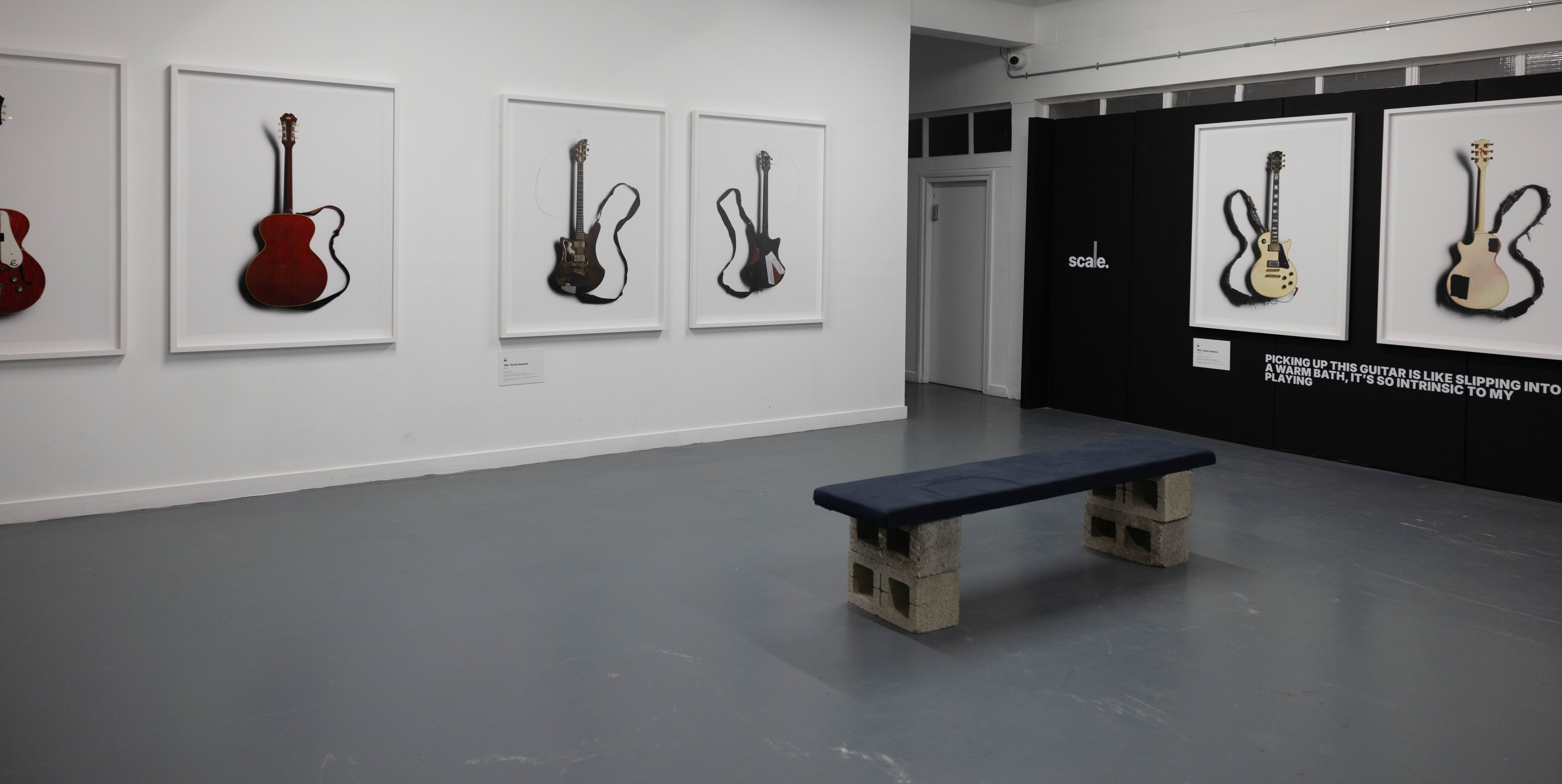 View of gallery space with bench in centre of room. White framed images of guitars are on the walls.