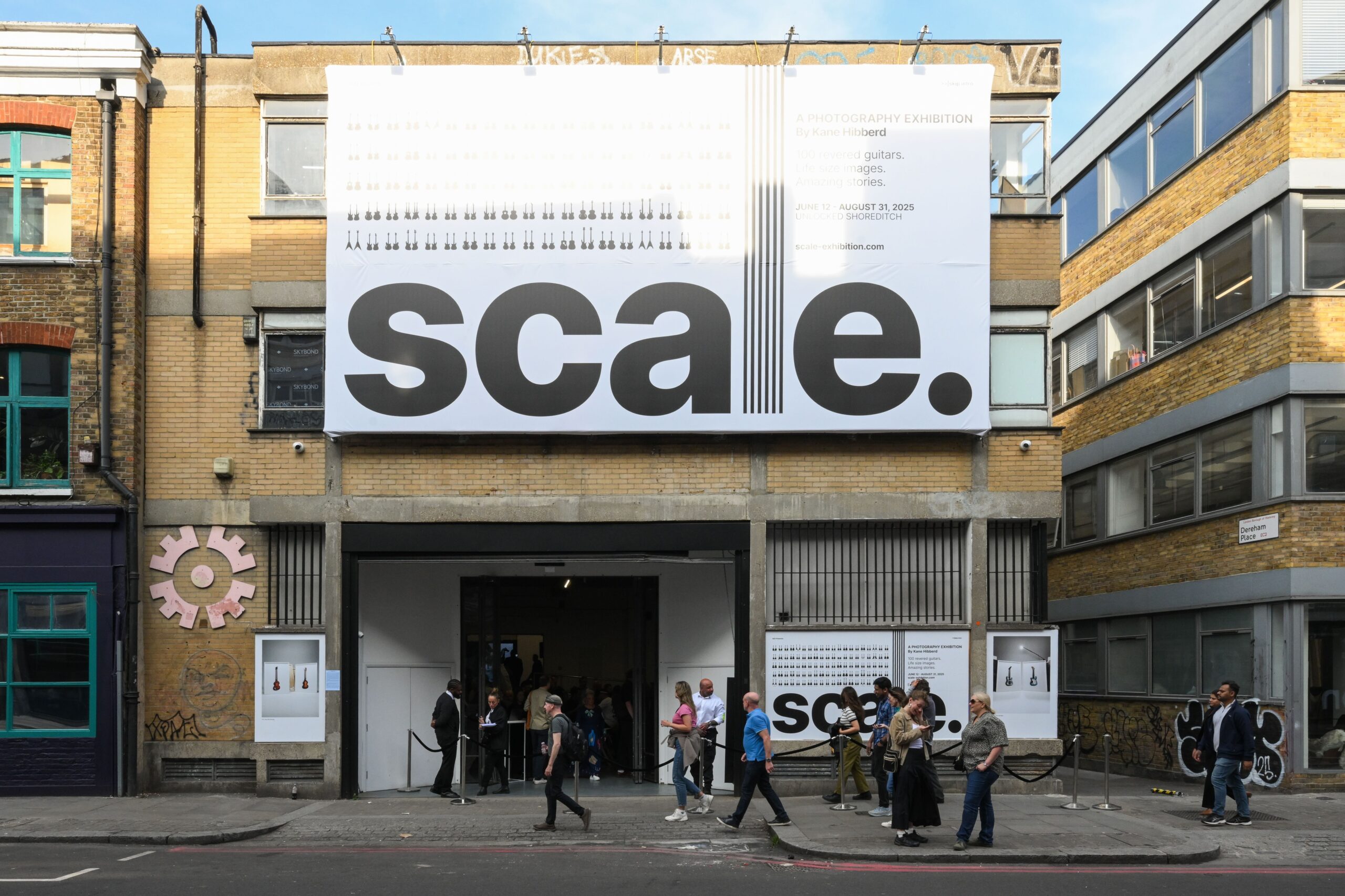 Front of yellow brick industrial building with people walking on street in front. Scale Exhibition billboard and signs are on front of building.
