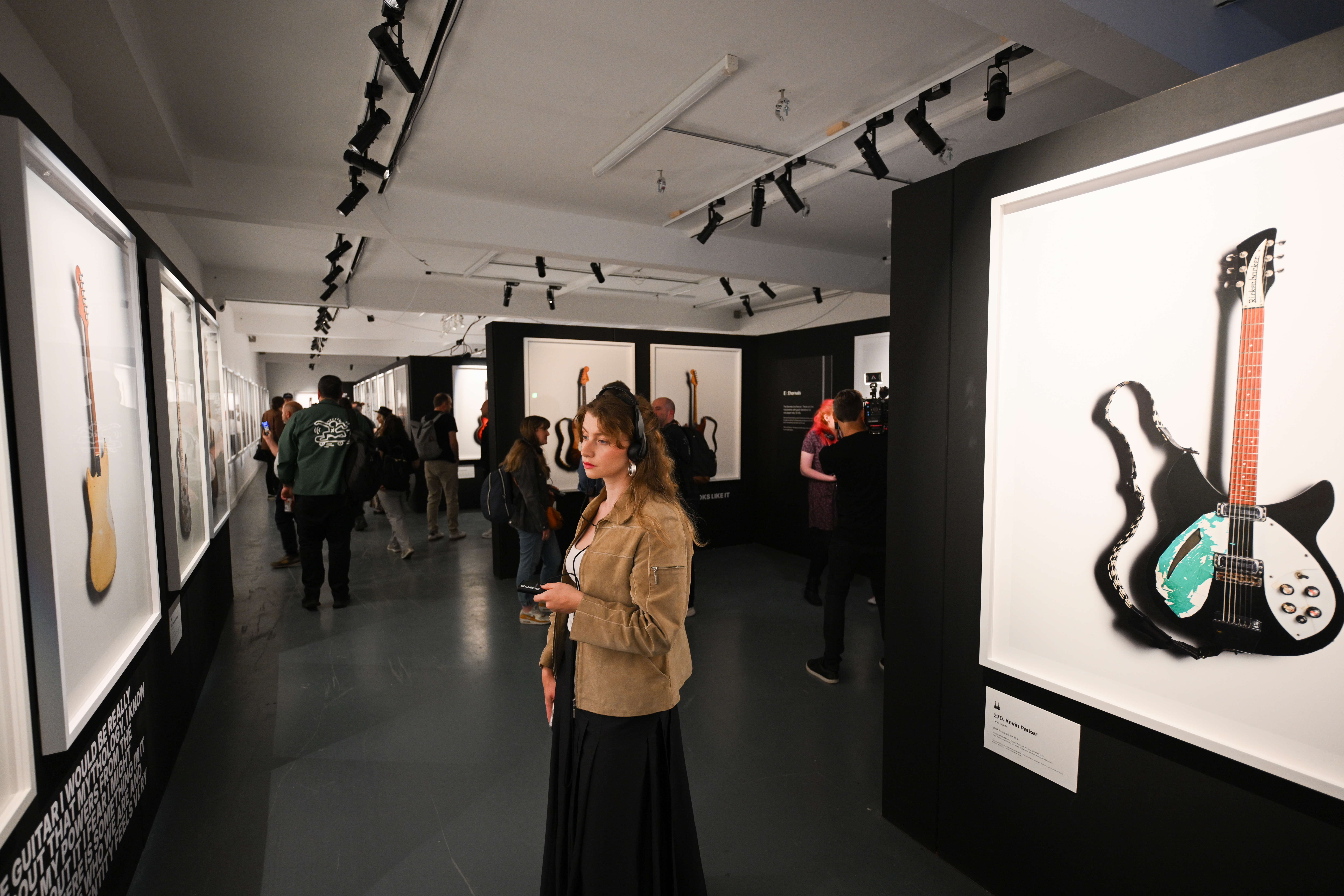 A woman stands in front of an image in a gallery wearing headphones. People can be seen looking at other images int he background.