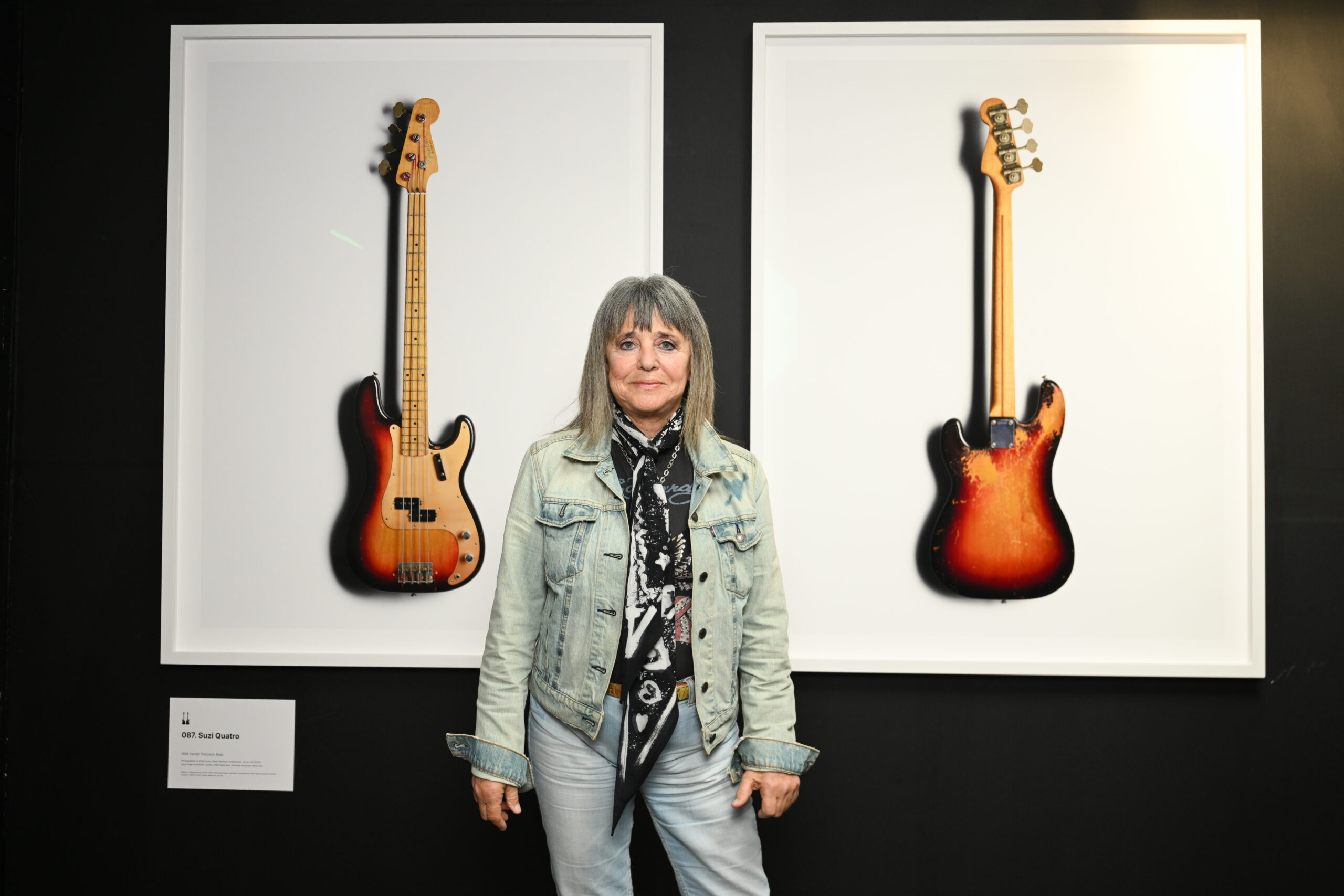 Musician Suzi Quatro stands in front of portraits of her guitar, front and back which are hung on a black wall.