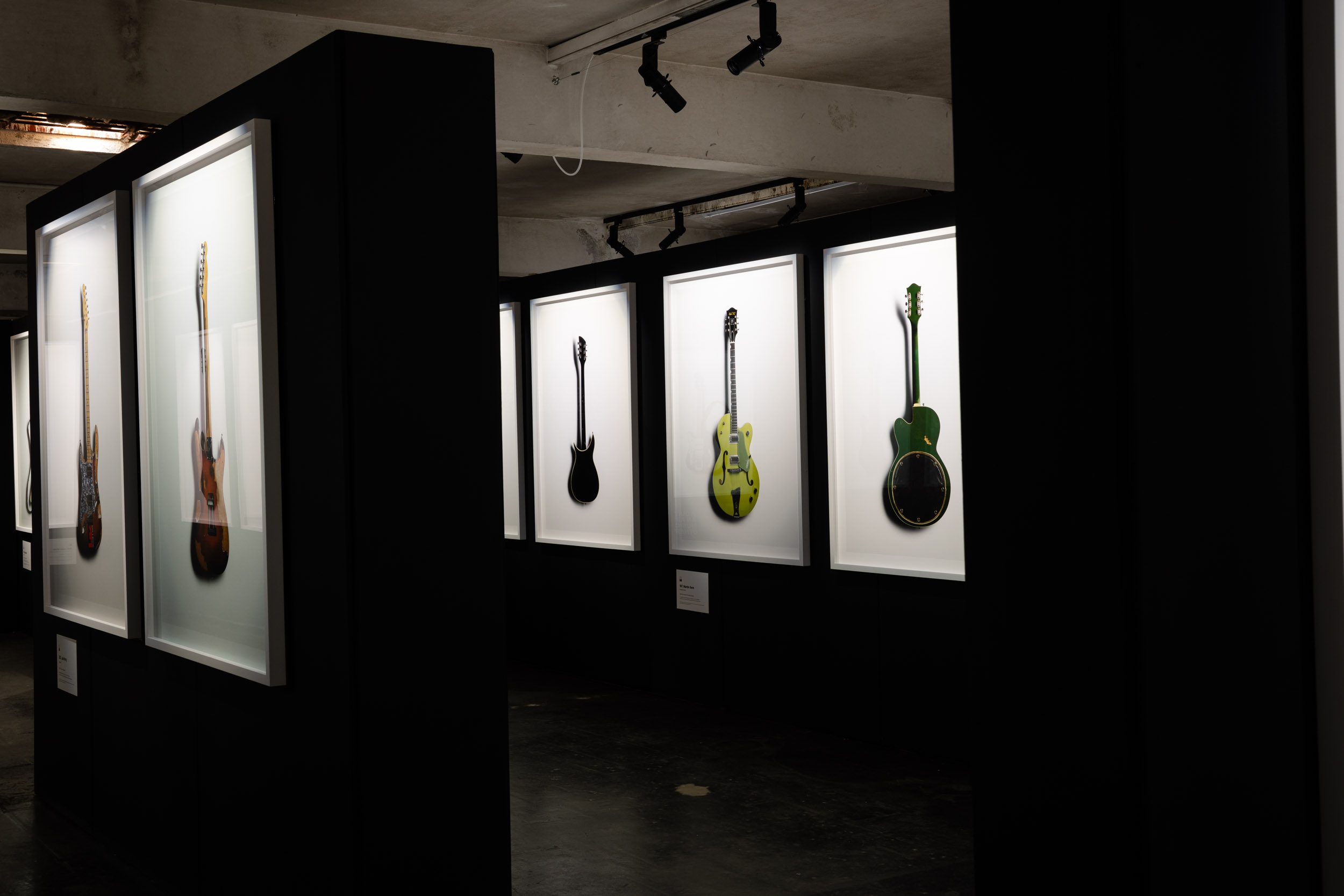 Life sized guitar images in white frames are hung on a black wall with down lighting